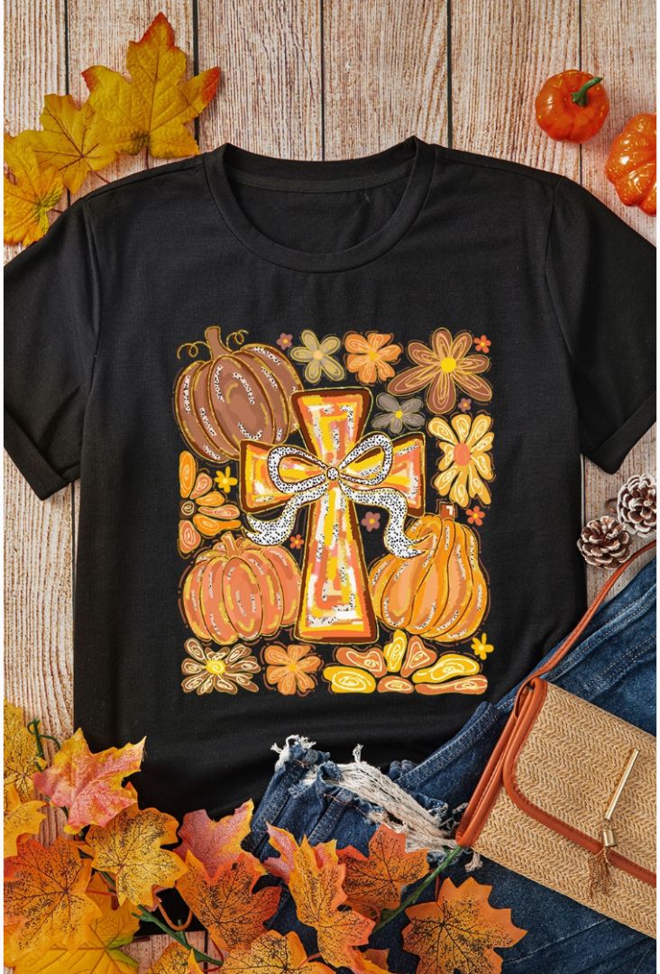 Black t-shirt with a cross and pumpkins design on a wooden surface with autumn leaves and a handbag.